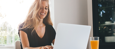 A smiling woman with long, blonde wavy hair is sitting at a table, looking at a silver laptop and typing. She is wearing a black shirt. A glass of orange juice is next to the laptop. A bright window and a stack of white plates are in the background.