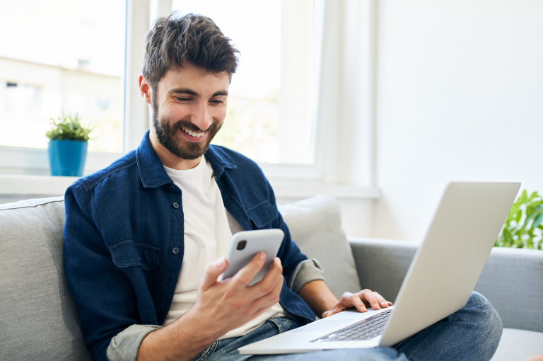 A man sitting on a couch, looking at his phone with a laptop beside him.