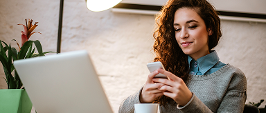 A woman smiling while looking at her phone, with a laptop in front of her.