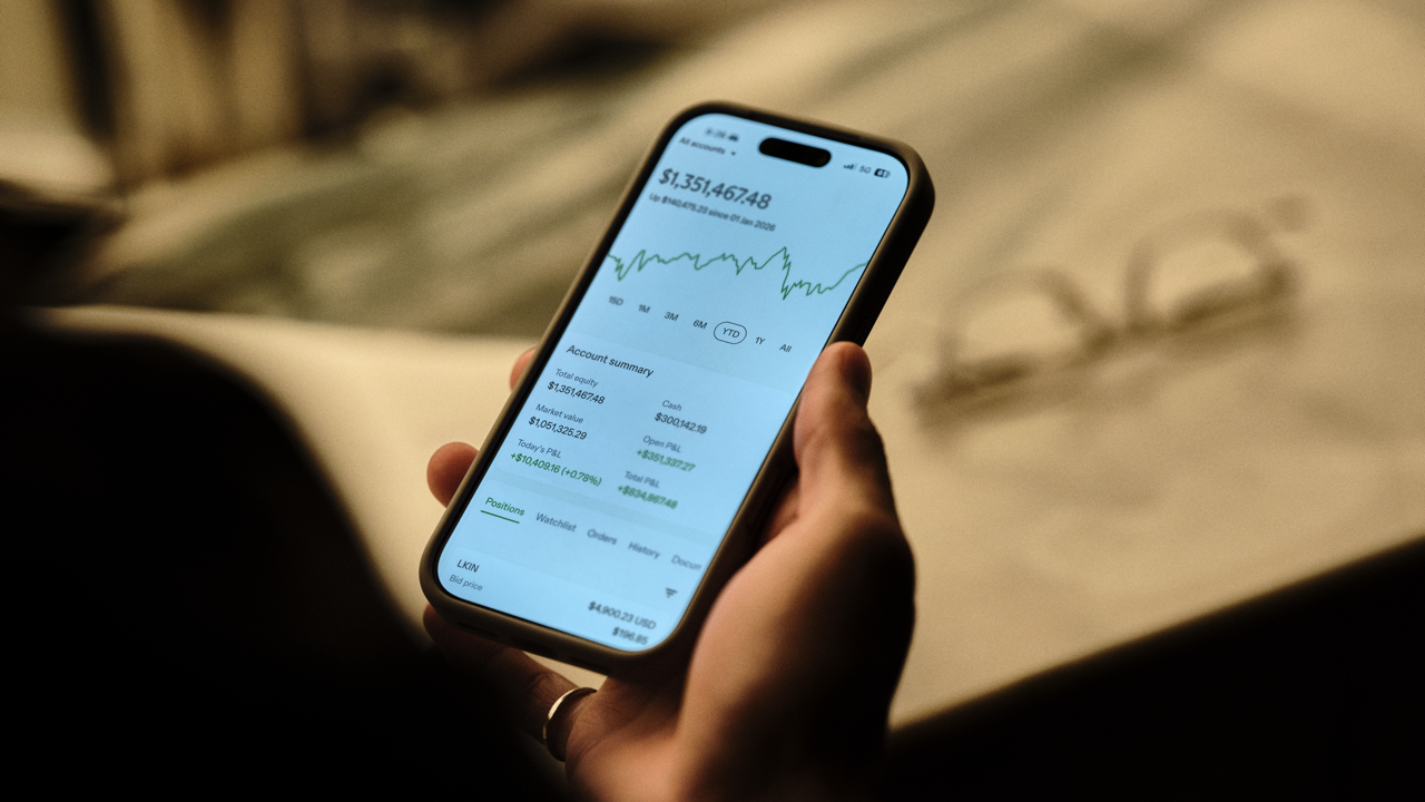 Close-up of a hand holding a phone showing an investment account summary with total equity, positive returns, and a year-to-date performance chart, glasses resting nearby in soft light.