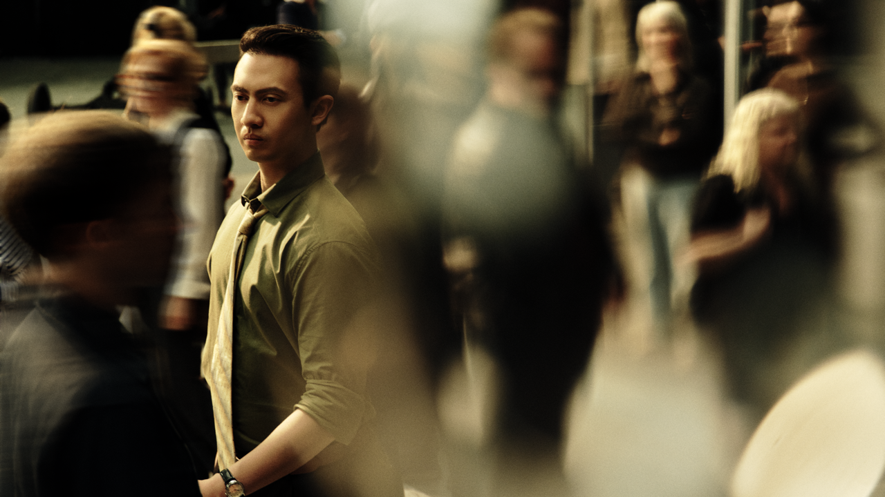 Man in a green shirt and tie standing still with a serious expression amid a crowd of blurred pedestrians in motion.