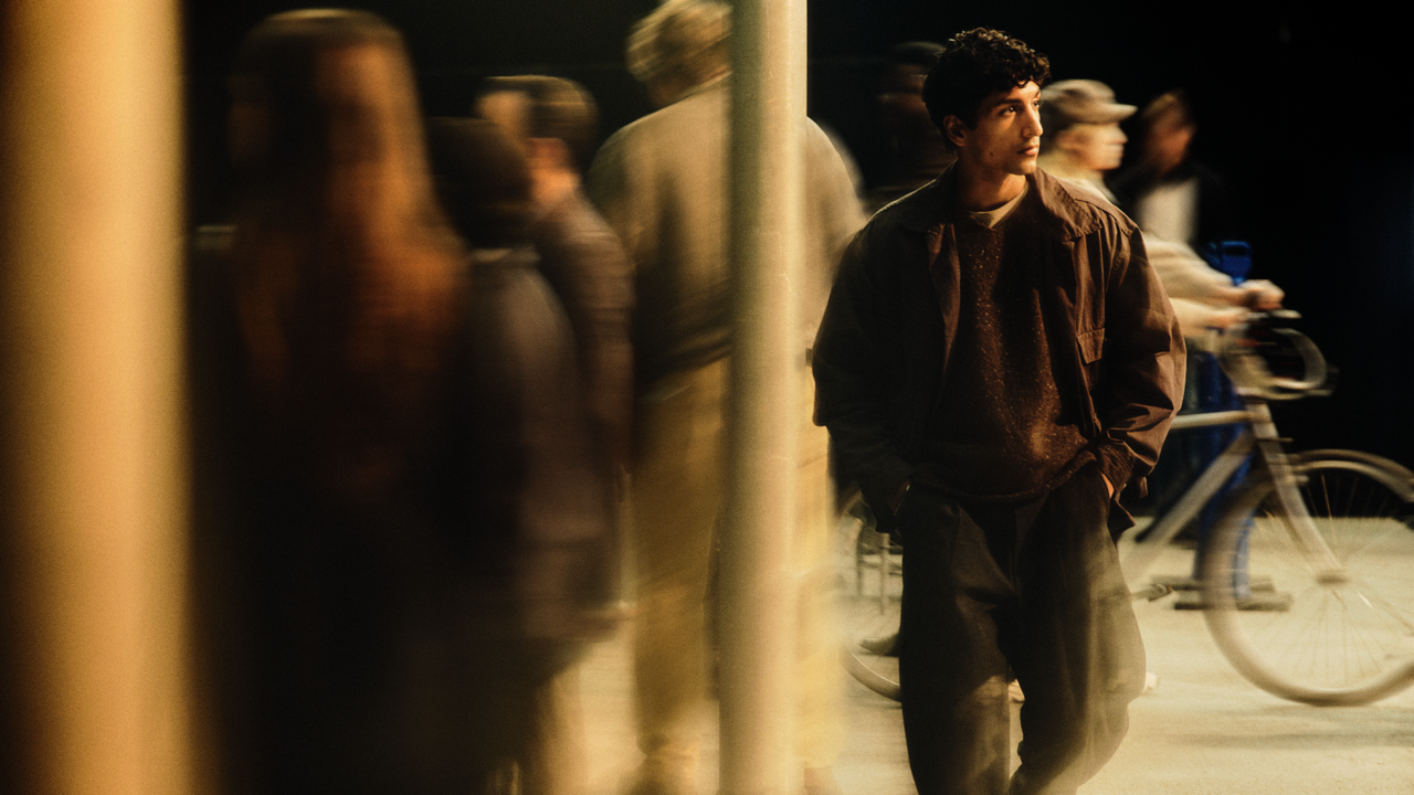 Young man with curly hair walking through a busy nighttime street, hands in pockets and looking to the side, with blurred pedestrians and a bicycle passing in the background under warm lighting.