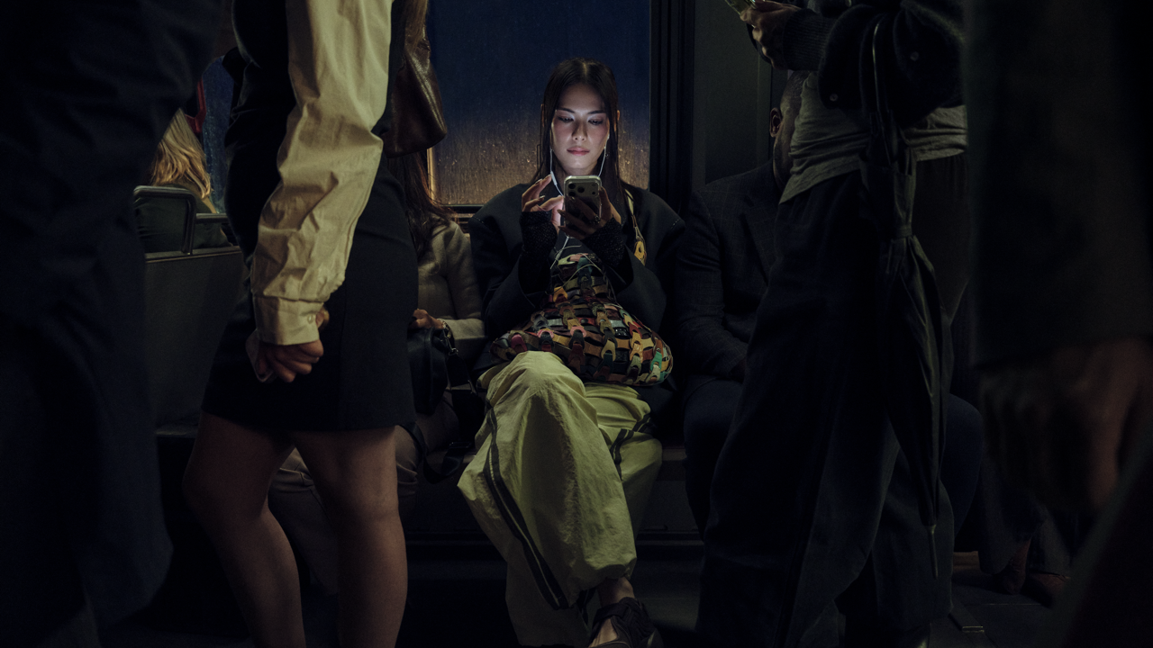 Woman seated on a crowded bus or train at night, illuminated by her phone screen while other passengers stand around her in shadow.