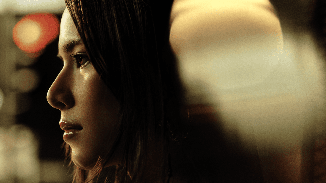 Close-up profile of a woman with long dark hair looking ahead with a serious expression, lit warmly against a blurred background with soft round light flares.