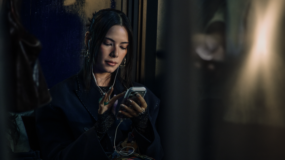 Woman with wet, dark hair and drop earrings sitting by a rain-streaked window, wearing earphones and looking down at her phone, lit softly by warm light against a dark interior.