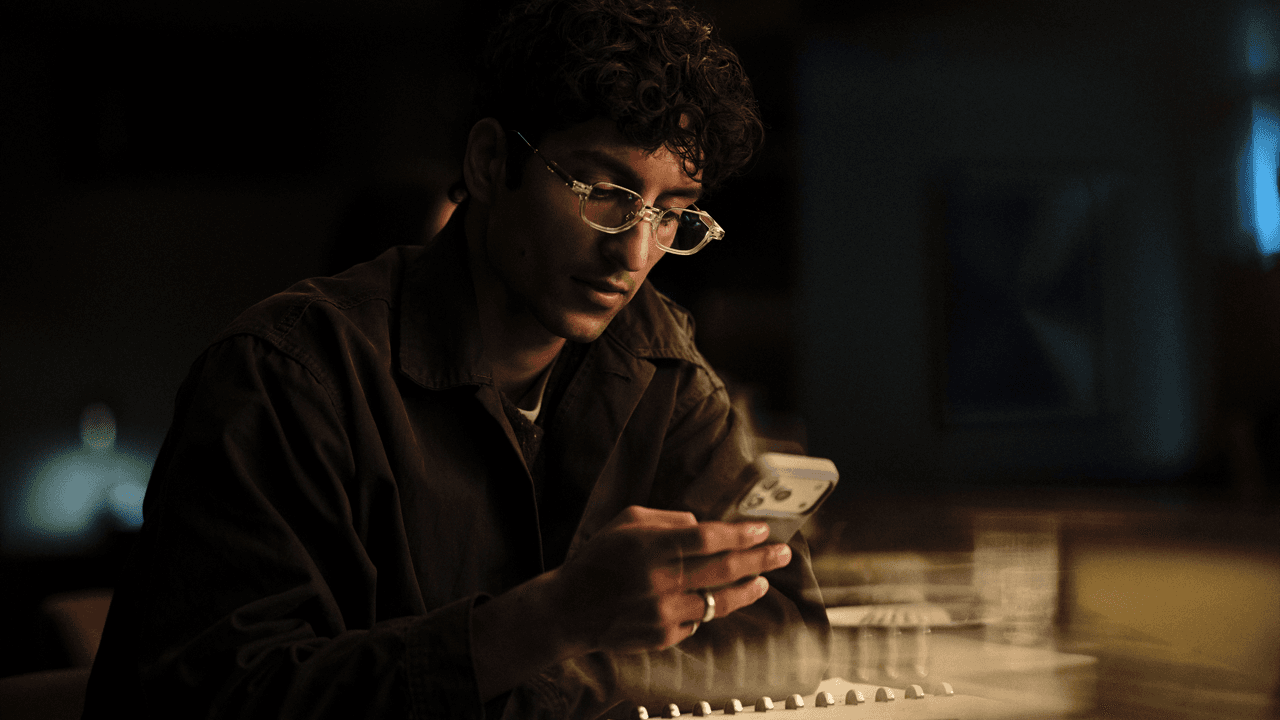 Man with curly hair and clear-framed glasses looking down at his phone, illuminated by its glow while seated at a table in a dimly lit room.