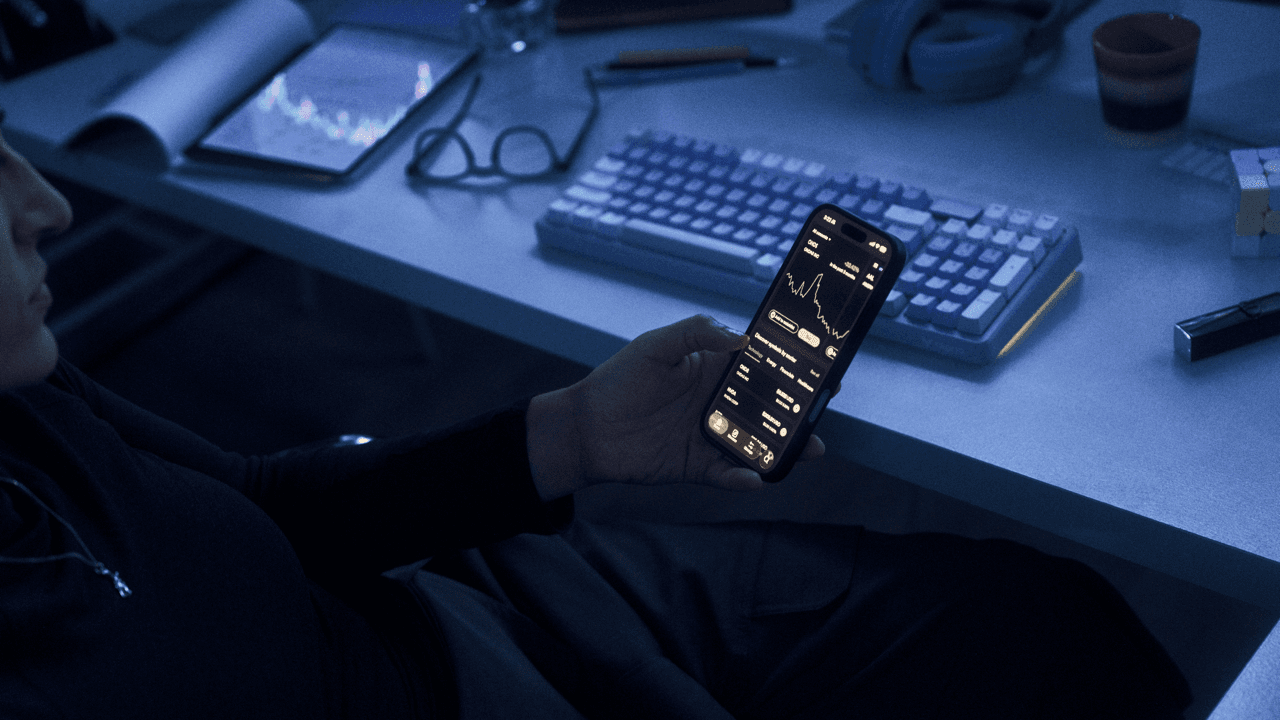 Person holding a phone displaying a stock trading app with charts and market data, at a dimly lit desk with a keyboard, tablet showing a price chart, glasses, and a Rubik's cube.